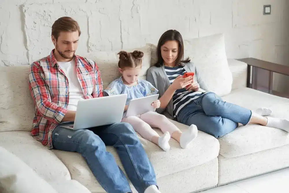 A co-parenting family sits together on a couch using laptop, tablet, and phone