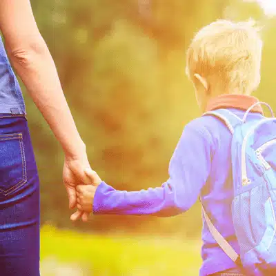 Parent holds child's hand while walking outdoors; backpack suggests school, custody, and caregiving responsibilities together
